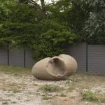 A sand sculpture with a large hole sits on gravel in front of a gray fence.