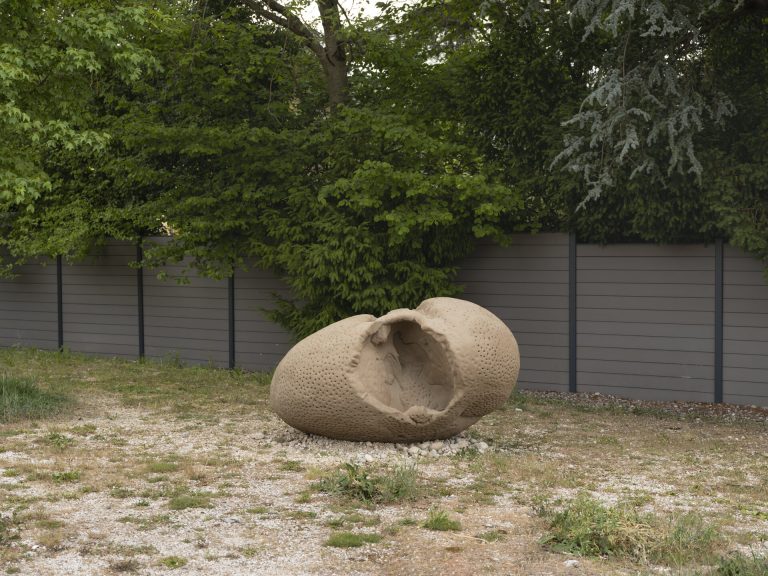 A sand sculpture with a large hole sits on gravel in front of a gray fence.