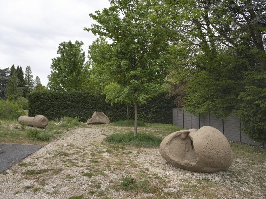 Three sand and resin sculptures sit on gravel near a hedge.