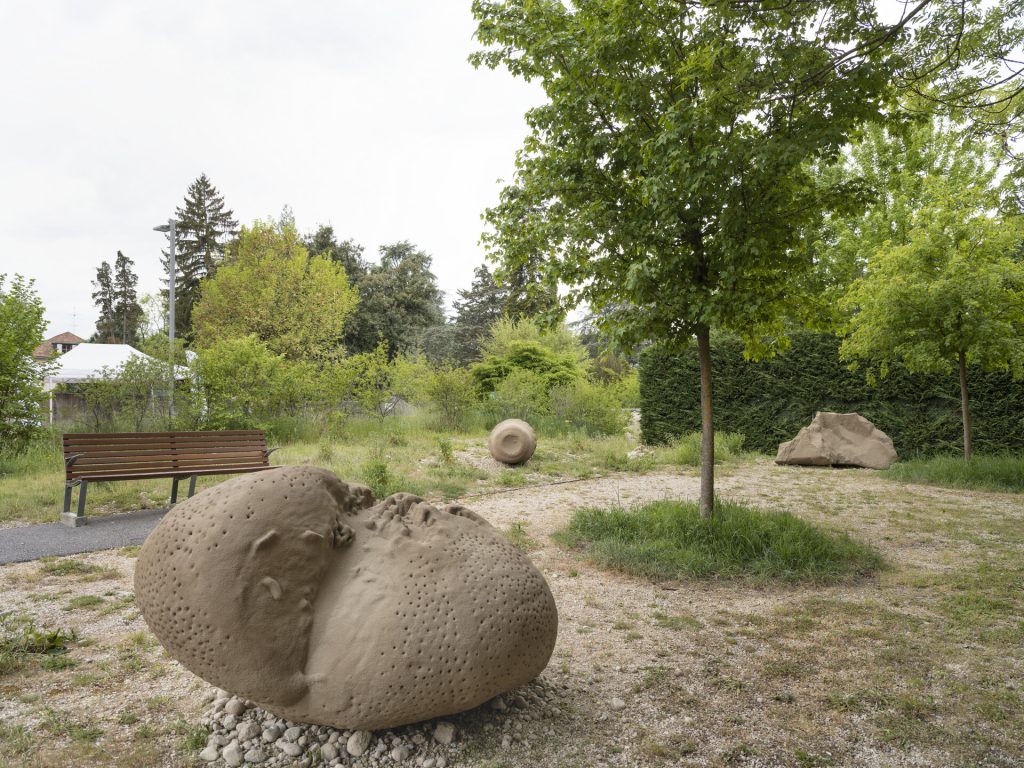 Three sand and resin sculptures sit on gravel in a park near a bench and trees.