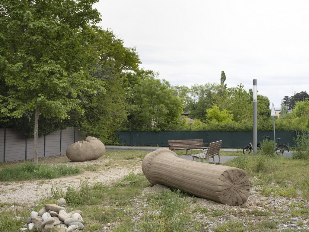 Two sand and resin sculptures sit on a gravel path near a bench and trees.