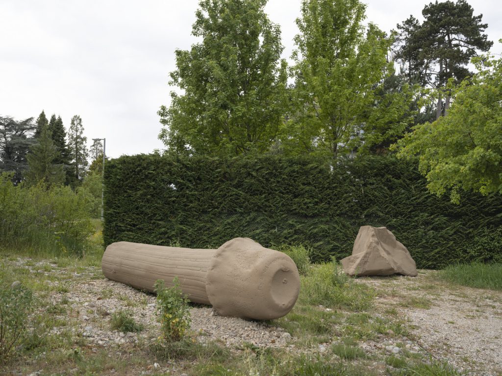 Two sand and resin sculptures sit on gravel before a green hedge.
