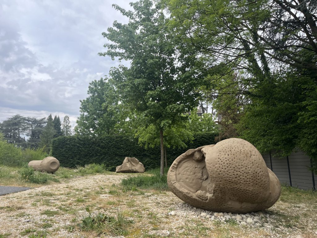Three sand, resin, and EPS sculptures sit on gravel and grass near trees.