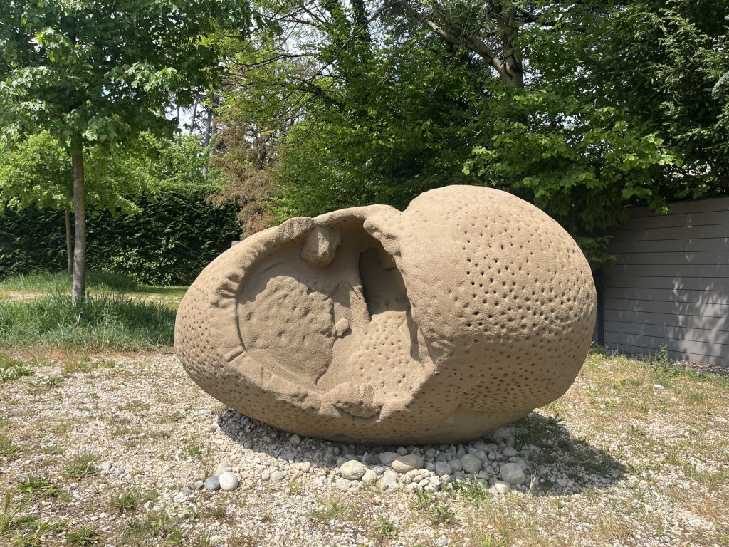 A sand and resin sculpture sits on gravel with a large opening and pitted surface.