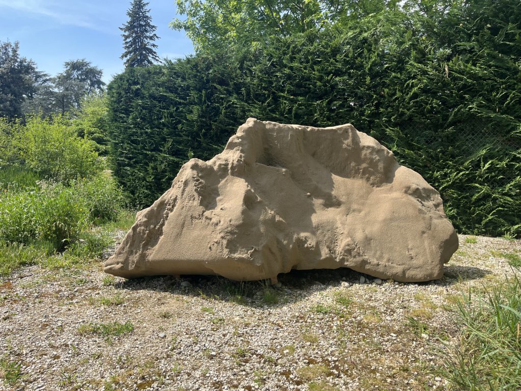 A sand sculpture sits on gravel in front of a green hedge.