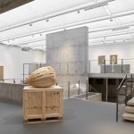 A 3d printed sand sculpture sits on a wooden crate in a gallery with concrete walls and stairs.