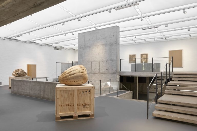 A 3d printed sand sculpture sits on a wooden crate in a gallery with concrete walls and stairs.