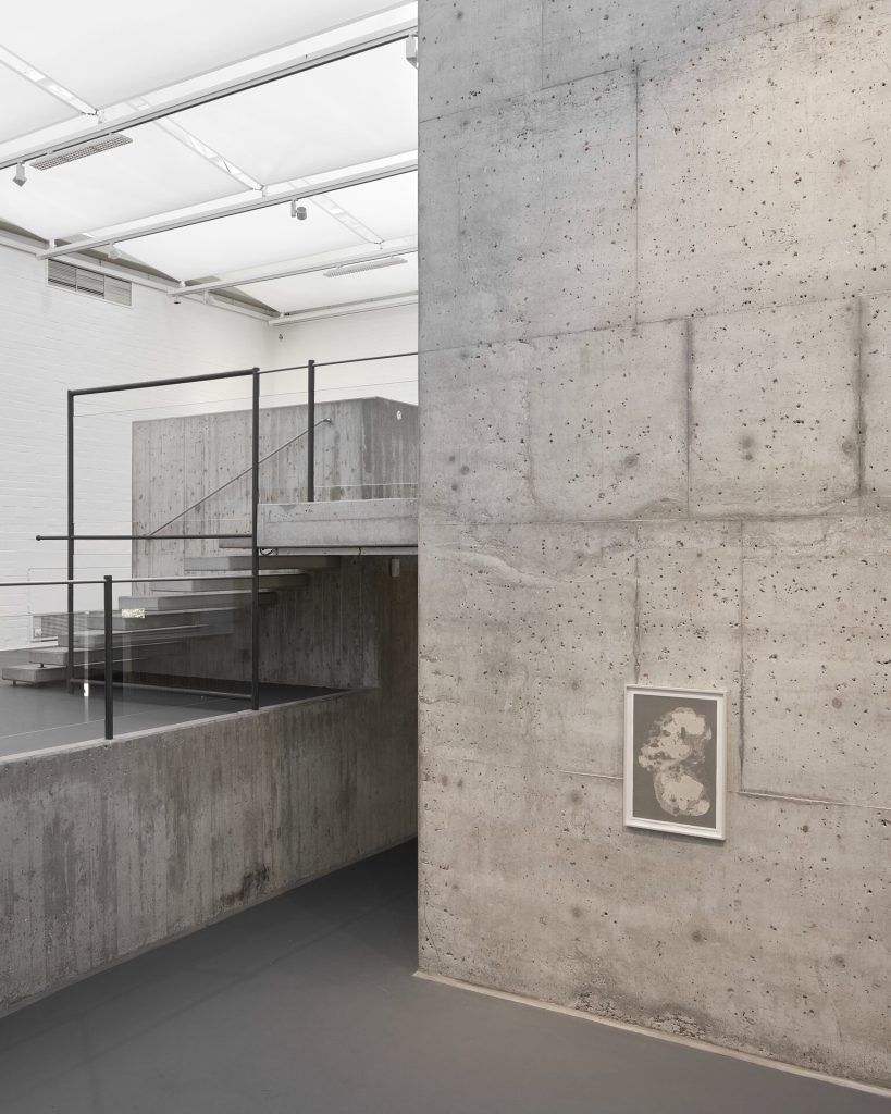 A framed screen print hangs on a concrete wall near a staircase with glass railings.