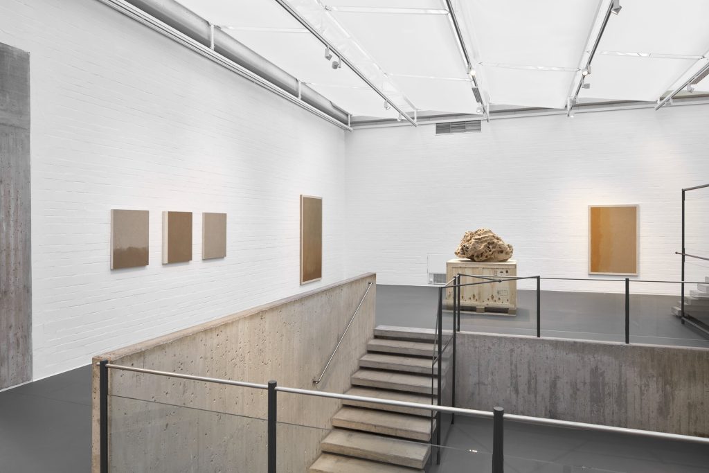 A 3d printed sand sculpture sits on a wooden crate above concrete stairs in a gallery with screenprints on white walls.