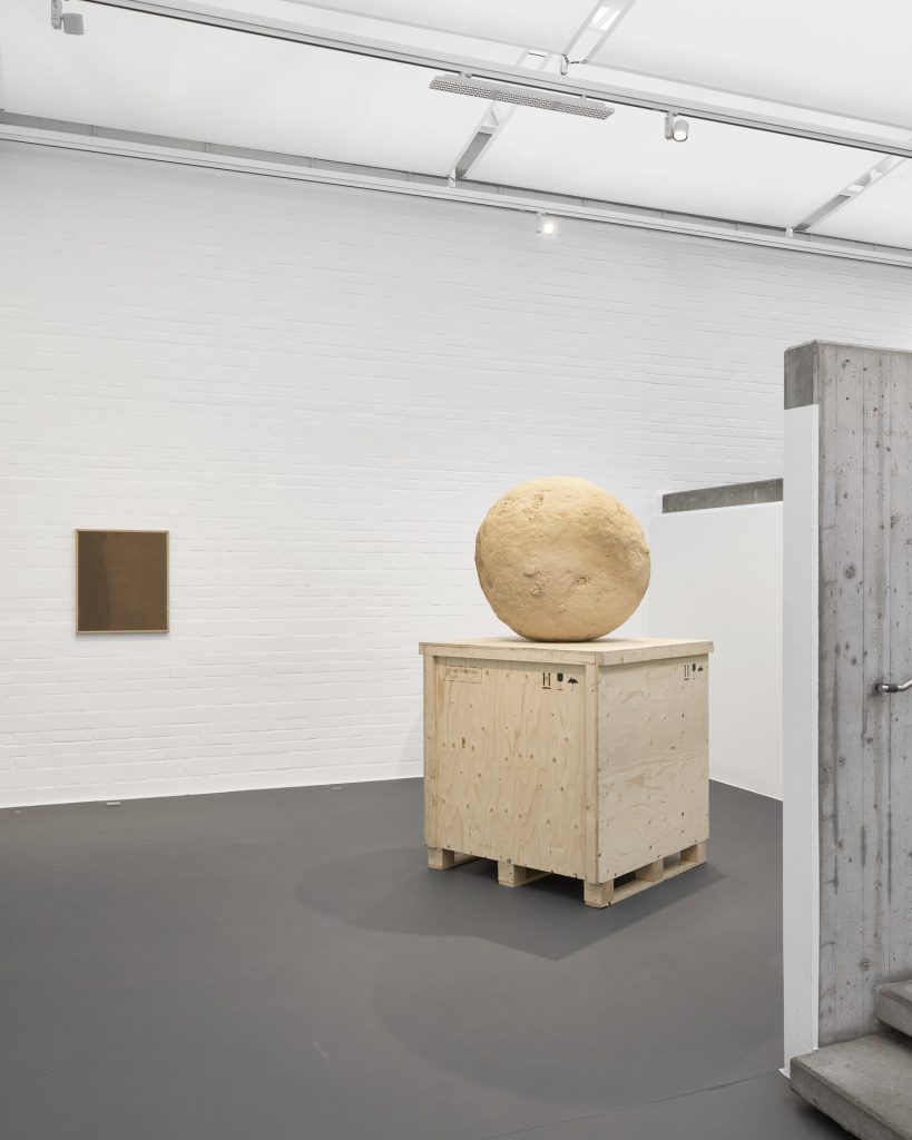 A 3d printed sand sculpture sphere sits on a wooden crate in a gallery.
