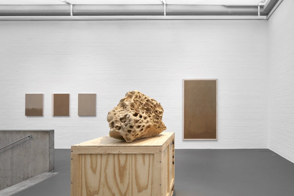 A porous 3d printed sand sculpture sits on a wooden crate in a gallery with screenprints on white walls.