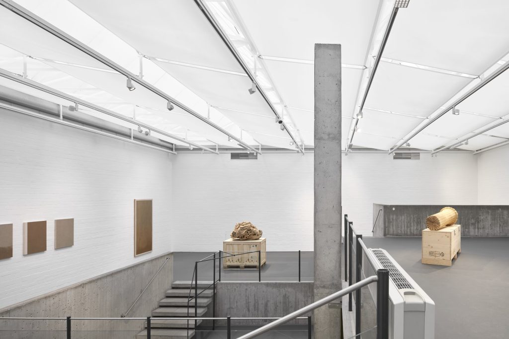 Sand sculptures rest on wooden crates in a white gallery with screenprints on the left wall.