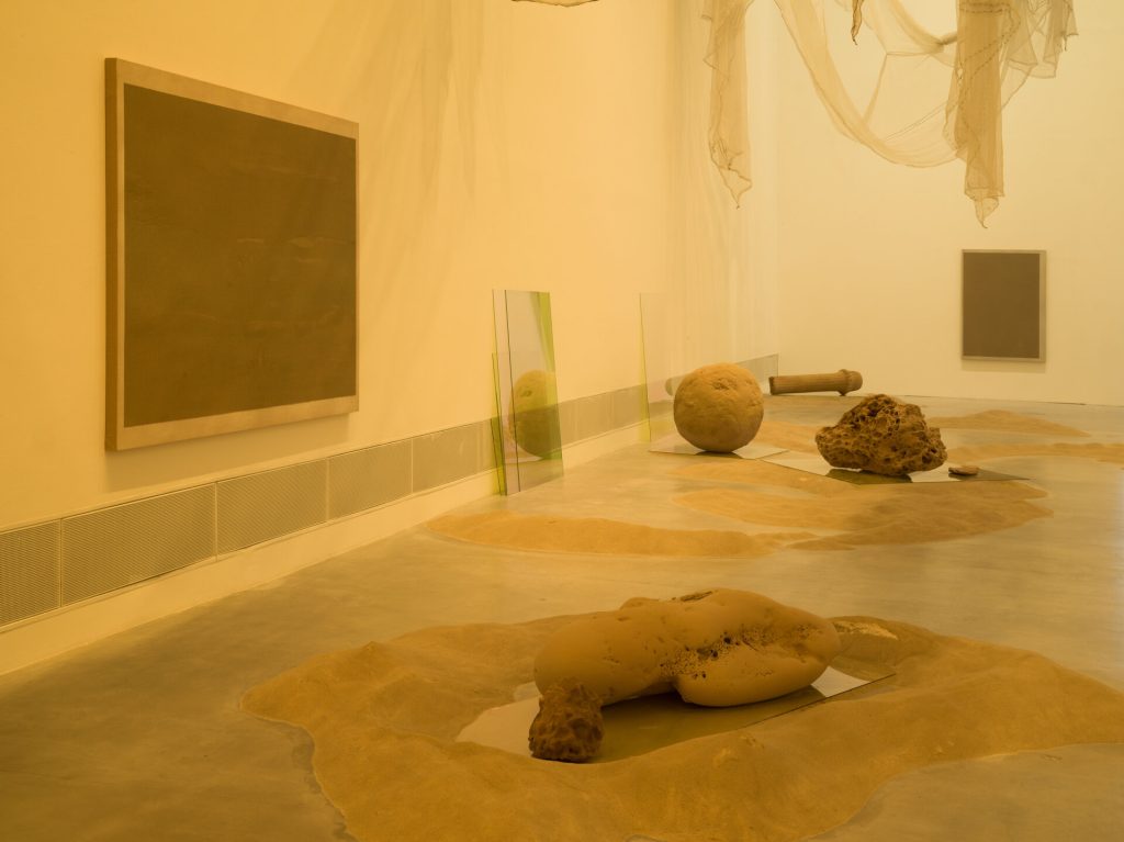 A gallery floor holds sand piles, glass panels, and a rounded sand form, with two framed sand paintings on the wall.