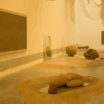 A gallery floor holds sand piles, glass panels, and a rounded sand form, with two framed sand paintings on the wall.