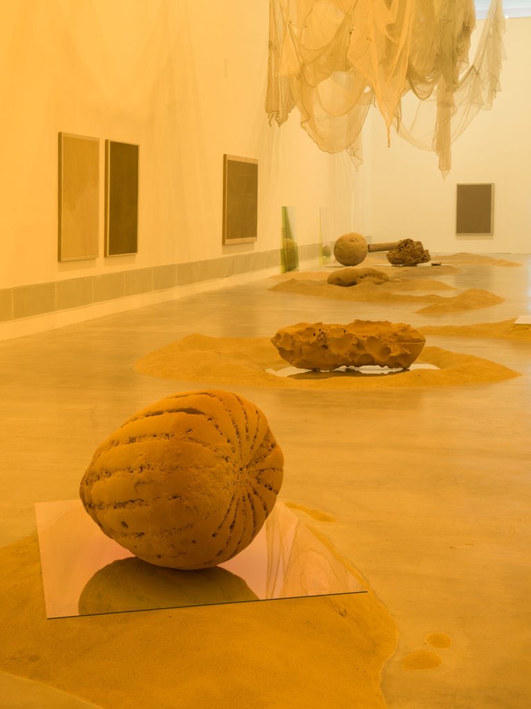 A 3D-printed sand sphere rests on a glass panel in a sand-covered gallery with sand paintings on the walls.