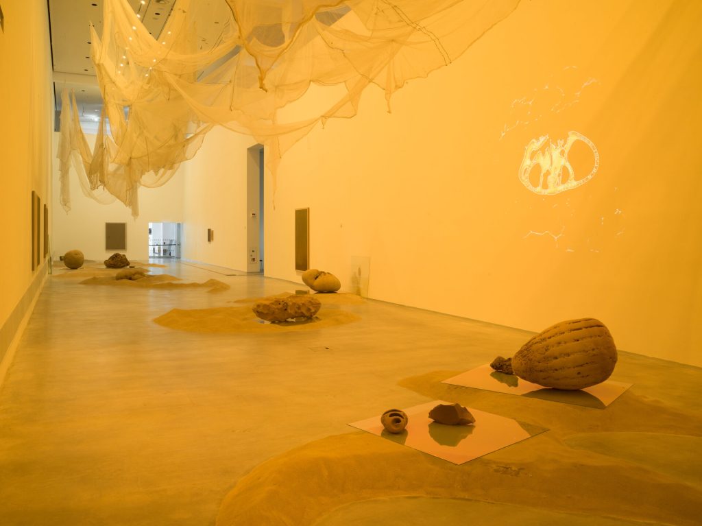 Sand mounds and rounded forms sit on glass panels in a yellow-lit gallery with hanging fabric.