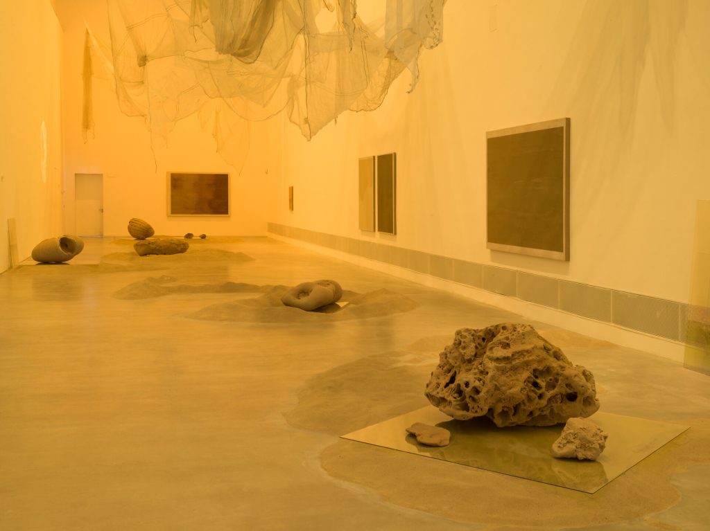 A gallery room lit in yellow holds sand mounds, porous 3D-printed sand sculptures, and framed sand paintings on the walls.