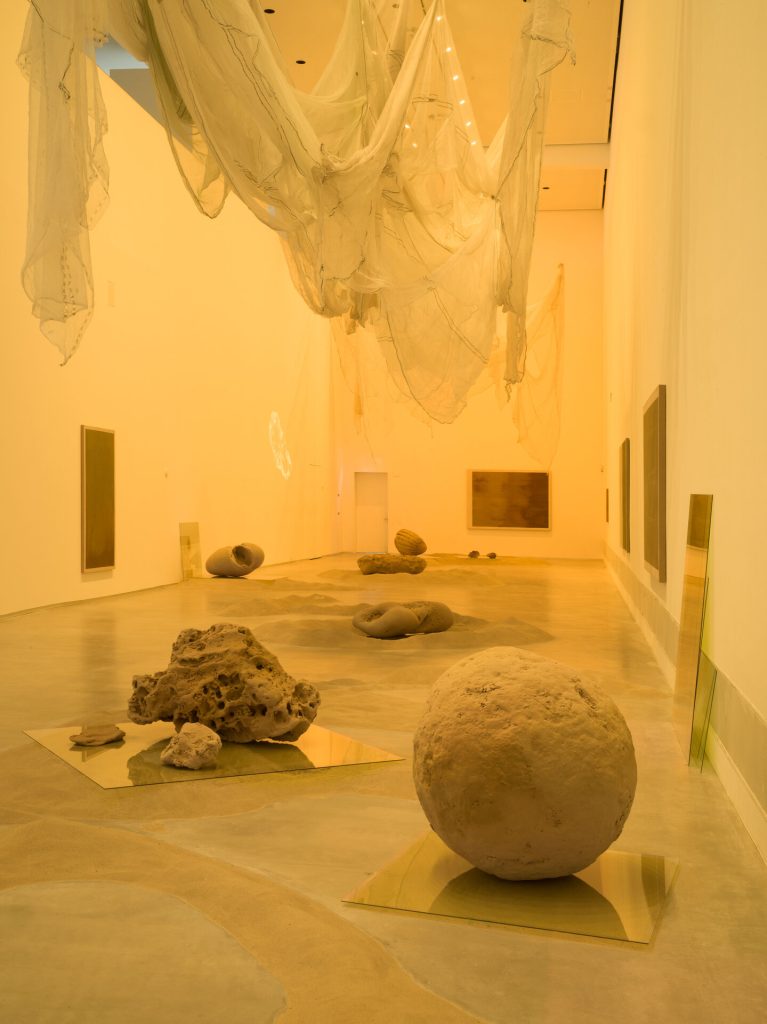 Sand sculptures sit on glass panels in a gallery, with sheer fabric hanging from the ceiling.