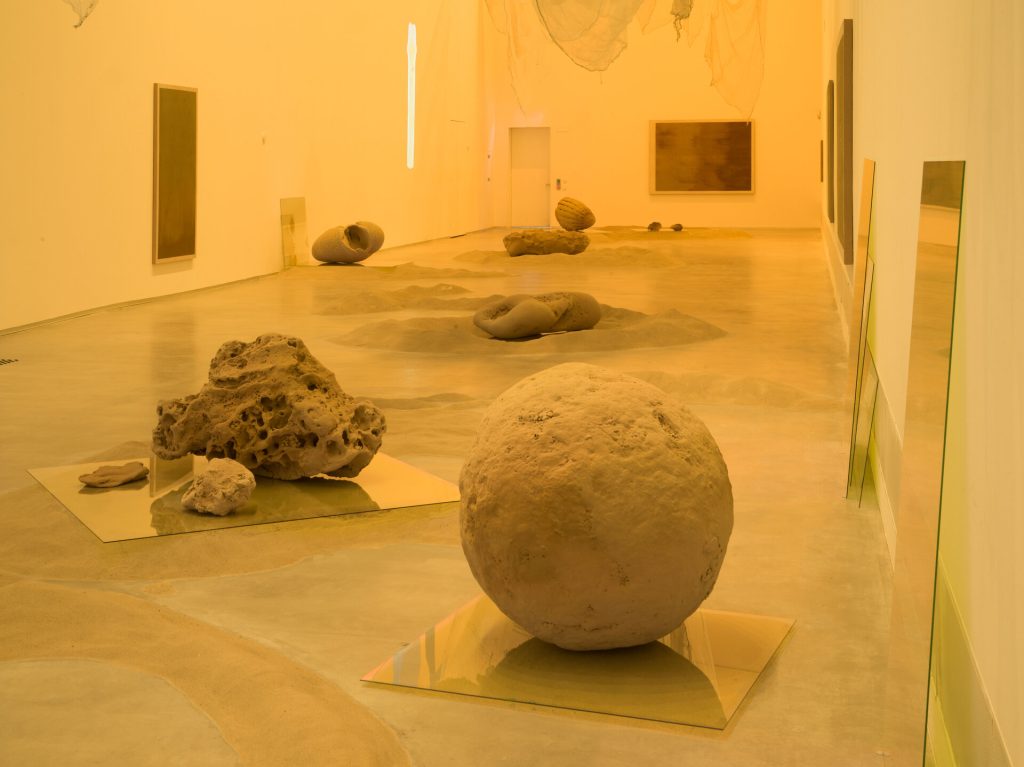 A gallery floor holds sand mounds and 3d printed sand sculptures on glass panels under yellow light.