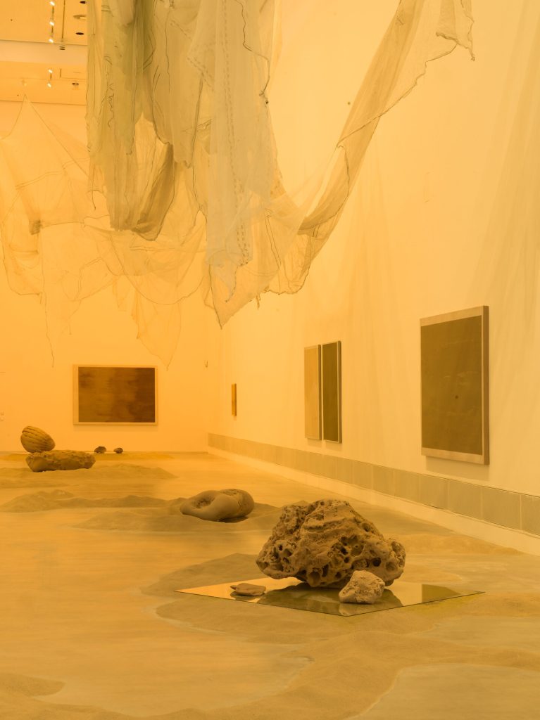 A gallery room with sand on the floor, a porous stone on a glass panel, and translucent fabric hanging from the ceiling.
